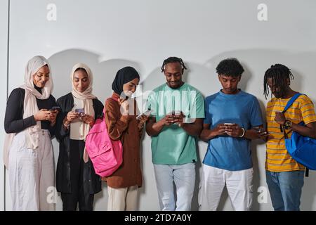Eine vielfältige Gruppe von Schülern, die in das digitale Zeitalter eingetaucht sind, steht vereint, während sie mit ihren Smartphones vor einem weißen Hintergrund interagieren und symbolisieren Stockfoto