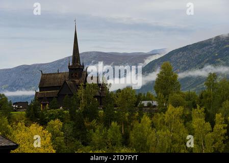 LOM Stave Church mit Bergen und niedrigen Wolken im Hintergrund und wechselnden Bäumen im Vordergrund. Stockfoto