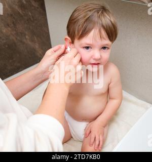 Eine Mutter reinigt ihre Kinderohren mit einem Wattepad im Badezimmer. Kind im Alter von zwei Jahren (zweijähriger Junge) Mama wäscht die Ohren des Kleinkindes Stockfoto