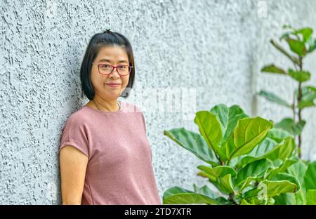 Porträt Asiatische Frau mittleren Alters, die im Garten an der Wand steht, Brille trägt, in die Kamera blickt, natürliches Licht, lächelnd, schwarz kurz h Stockfoto