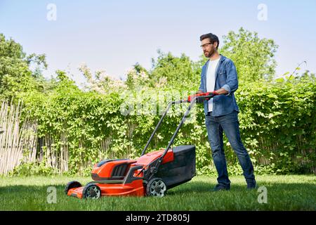 Schöner männlicher Gärtner in Handschuhen, der tagsüber Rasen mit einem elektrischen Mäher im Garten trimmt. Seitenansicht eines lächelnden bärtigen Mannes in Denim-Outfit mit Rasenbesatz Stockfoto