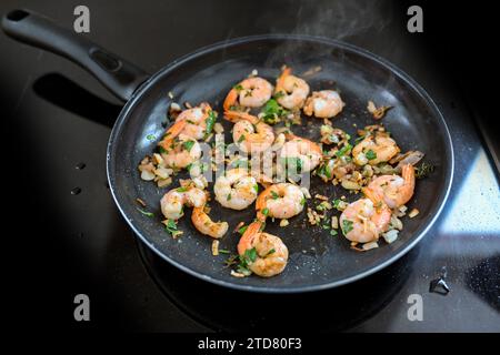 Garnelen mit Schwänzen werden in einer Pfanne mit Zwiebeln, Knoblauch und Kräutern auf einem schwarzen Herd geröstet, eine Meeresfrüchtemahlzeit kochen, einen Kopierraum, einen ausgewählten Fokus, Stockfoto