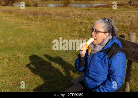 Seniorenwanderer essen Bananen, machen eine Pause auf Holzbank, genießen sonnigen Herbsttag im Naturschutzgebiet, kleiner Teich im Hintergrund, Winte Stockfoto