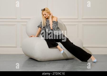 Eine schöne blonde Frau in modischer Kleidung im Boho-Stil sitzt mit überkreuzten Beinen in einem weichen Sitzsack-Stuhl. Ihre lose Hose und Stiletto Stockfoto