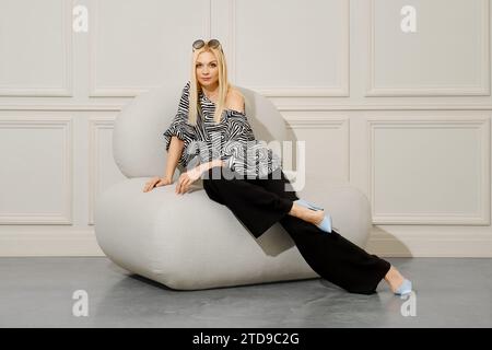Eine modische Frau sitzt in einem weichen Stuhl und trägt eine breite Hose und eine gemusterte Bluse. Ihr blondes Haar ist perfekt gestylt und sie trägt eine Sonnenbrille Stockfoto