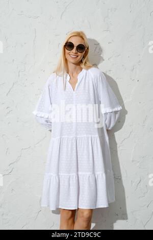 Zufriedene blonde Frau in Sonnenbrille und weißem Sommerkleid, das an die Wand gedrückt wurde Stockfoto