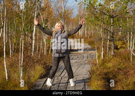 Eine glückliche Frau mit Rucksack springt auf einem Holzweg in einem Sumpf in Jelnya, Weißrussland. Stockfoto