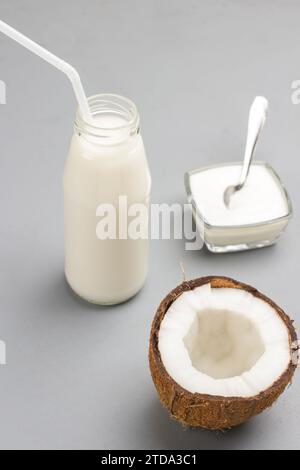 Halb frische Kokosnuss. Kokosmilchflasche, Strohhalme. Schüssel Joghurt, Löffel. Grauer Hintergrund Stockfoto