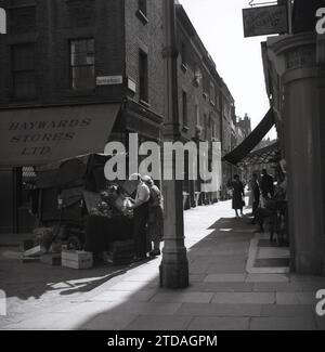 1950er Jahre, historisch, eine Käuferin mit einem männlichen Händler am Marktstand, Shepherd Market, in Mayfair, London, England, UK. An der Ecke der gepflasterten Gasse, Hayward Stores und ein Schild für gute Wohnungswirtschaft Magazin für 1,9m im Monat. Der Shepherd Market liegt in Mayfair in der Nähe von Piccadilly und ist ein bezaubernder Teil des Zentrums von London mit seinem kleinen Platz und engen Seitenstraßen, bekannt als „Dorf in Piccadilly“. Stockfoto