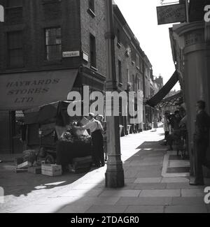 1950er Jahre, historisch, eine Käuferin mit einem männlichen Händler am Marktstand, Shepherd Market, in Mayfair, London, England, UK. An der Ecke der gepflasterten Gasse, Hayward Stores und ein Schild für gute Wohnungswirtschaft Magazin für 1,9m im Monat. Der Shepherd Market liegt in Mayfair in der Nähe von Piccadilly und ist ein bezaubernder Teil des Zentrums von London mit seinem kleinen Platz und engen Seitenstraßen, bekannt als „Dorf in Piccadilly“. Stockfoto