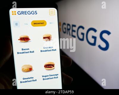 Person mit Smartphone und Webseite der britischen Bäckereikette Greggs plc vor dem Geschäftslogo. Konzentrieren Sie sich auf die Mitte des Telefondisplays. Stockfoto