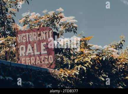 Bellagio Italien - 8. Mai 2011; altes, rostiges und beschädigtes Restaurantschild auf dem Dach, umgeben von blühenden Bäumen in der italienischen Touristenstadt Stockfoto