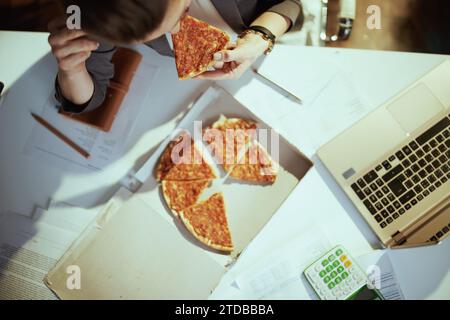 Nachhaltiger Arbeitsplatz. Obersicht einer Arbeitnehmerin im grünen Büro mit Pizza und Laptop. Stockfoto