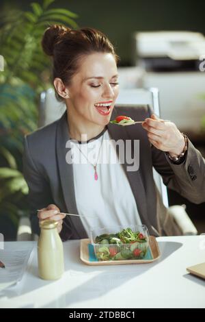 Nachhaltiger Arbeitsplatz. Glückliche moderne 40-jährige Angestellte in einem grauen Business-Anzug im modernen grünen Büro, die Salat isst. Stockfoto