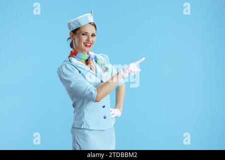 Lächelnde, stilvolle Stewardess-Frau auf blauem Hintergrund in blauer Uniform, die auf etwas zeigt. Stockfoto