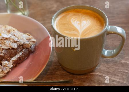 Ein Latte in einer Kaffeetasse, der morgens in einem Café neben einem Mandelcroissant getrunken wird. Herzsymbol in der Creme auf dem Kaffee. UK Stockfoto