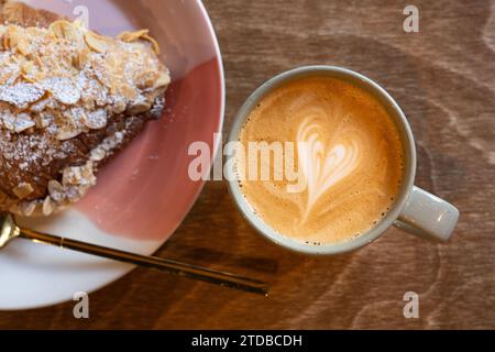 Ein Latte in einer Kaffeetasse, der morgens in einem Café neben einem Mandelcroissant getrunken wird. Herzsymbol in der Creme auf dem Kaffee. UK Stockfoto