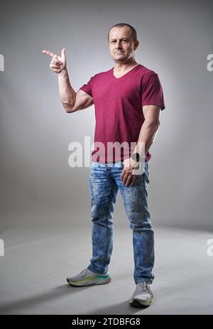 Studioporträt eines Mannes mittleren Alters in T-Shirt und Jeans auf grauem Hintergrund, der auf etwas seitlich des Bildes zeigt Stockfoto