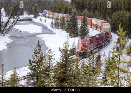 Containerzug, der von einer leistungsstarken Diesellokomotive auf einer Windlaufbahn gezogen wird, die an einem schneebedeckten Wintertag entlang eines gefrorenen Flusses in den Kanadischen Rockies fährt Stockfoto