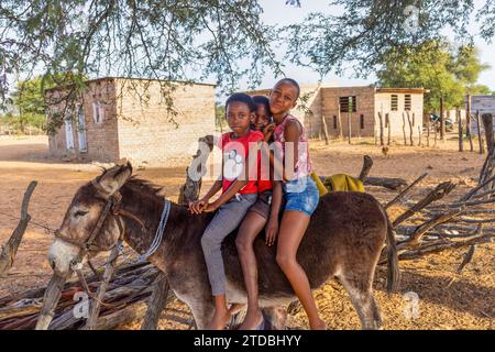 Dorf, afrikanische Kinder, die auf dem Bauernhof auf einem Esel reiten, Township Häuser im Hintergrund Stockfoto