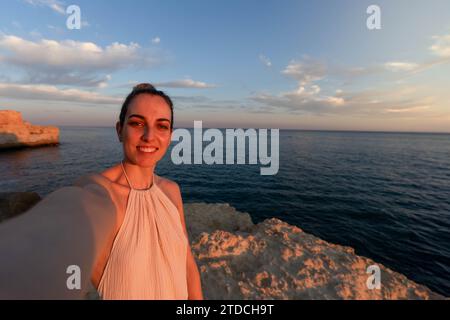 Serene Solitude: Ein Nahaufnahme, Selfie einer jungen Frau in einem Kleid, von oben auf einer Felsenklippe mit Blick auf den Ozean bei Sonnenuntergang aufgenommen. Stockfoto