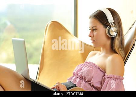 Profil einer Frau, die zu Hause Laptop und Kopfhörer auf einem Stuhl benutzt Stockfoto