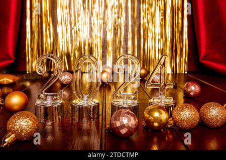 Auf hölzernem braunen Hintergrund transparente Zahlen 2024 unter Weihnachtsdekorationen Kugeln auf Textur von Folie goldenem Lametta, burgunderfarbenem Stoff. Symbiss Neujahr Stockfoto