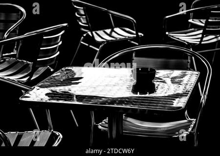 Minimales Schwarz-weiß-Bild eines Metalltisches umgeben von Stühlen. Schwarzer Aschenbecher auf Aluminiumoberfläche. Stockfoto