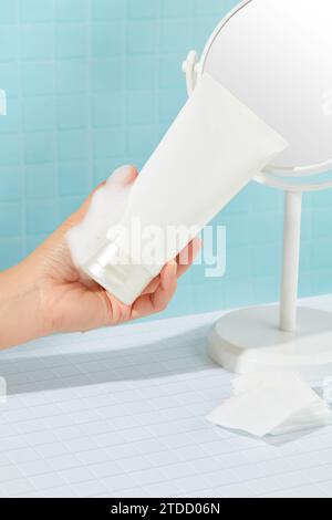 Eine Hand mit einer Tube Gesichtsreiniger ohne Marke mit weißem Schaum, einem Spiegel und Wattepads auf blauem und weißem Hintergrund. Kopierbereich für Kosmetik Stockfoto