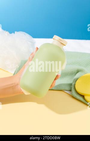 Nahaufnahme einer Hand mit weißem Schaum, die eine unbeschriftete Flasche Duschgel neben einem Handtuch, einem Badeschwamm und einem Schwamm hält. Bad mit Körper Stockfoto
