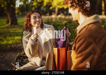 Ein Paar sitzt auf einer Bank im Park. Die Frau zeigt ihrem Mann nach dem Einkaufen Kleidung. Stockfoto
