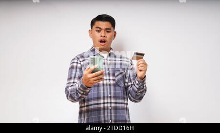 Asiatischer junger Mann mit schockiertem Ausdruck, der Kreditkarte und Smartphone hält, mit großen Augen auf den Smartphone-Bildschirm blickt, Innenstudio-Aufnahme isoliert Stockfoto
