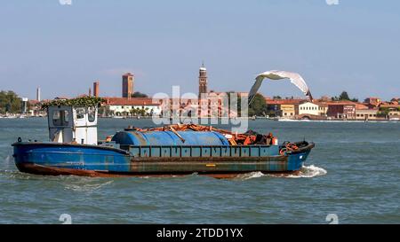 Eine Möwe fliegt über ein altes Schiff mit einem Kran in der Lagune von Venedig, Italien, mit der Insel Murano im Hintergrund Stockfoto