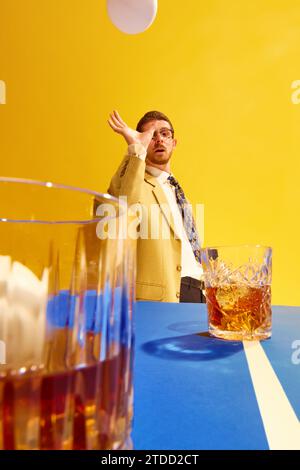 Junger Mann, Geschäftsmann in stilvollem Anzug, der Tischtennis bezahlt, Ball in Whiskey-Glas vor hellgelbem Hintergrund zielt Stockfoto