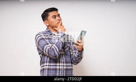 Junger asiatischer Mann, der sein Handy mit traurigem und verwirrtem Ausdruck auf weißem Hintergrund hält. studio-Aufnahme Stockfoto