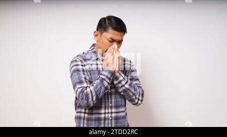 Junger erwachsener asiatischer Mann bedeckt Nase und Mund mit Gewebe, wenn er auf weißem Hintergrund niest. studio-Aufnahme Stockfoto