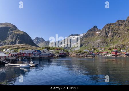 Panoramablick vom Wasser der klassischen roten Holzhäuser (Rorbu) an den felsigen Ufern von Moskenes, umgeben von Bergen auf den Lofoten Inseln Stockfoto