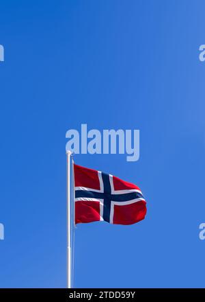 Vertikales Foto einer leuchtend flatternden norwegischen Flagge auf einem Fahnenmast vor einem klaren blauen Himmel Stockfoto