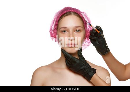 Porträt einer Frau mittleren Alters in rosa Kappen für kosmetische Behandlungen bei Schönheitsinjektionen. Hände in schwarzen Handschuhen halten die Spritze. Facelift. Stockfoto