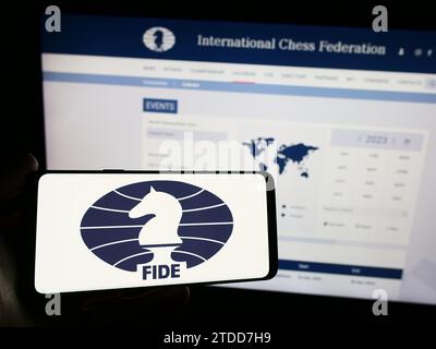 Person mit Smartphone und Logo der Organisation International Chess Federation (FIDE) vor der Website. Schwerpunkt auf dem Display des Telefons. Stockfoto