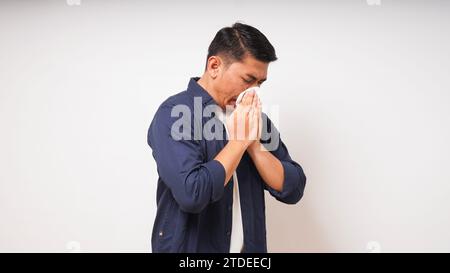 Junger erwachsener asiatischer Mann bedeckt Nase und Mund mit Gewebe, wenn er auf weißem Hintergrund niest. studio-Aufnahme Stockfoto