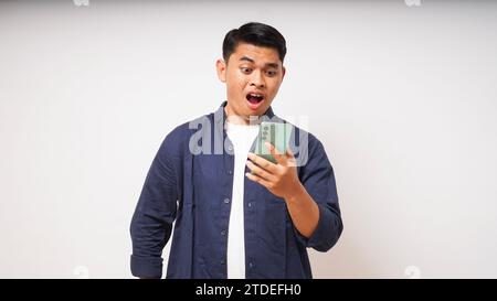 Junger asiatischer Mann mit schockiertem Gesichtsausdruck, während er ein Mobiltelefon auf weißem Hintergrund benutzt Stockfoto