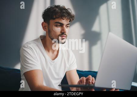 Porträt eines attraktiven bärtigen Mannes, der legere Kleidung trägt und auf einer Couch im Wohnzimmer sitzt und einen Laptop benutzt. Im Internet surfen. Verwenden Stockfoto