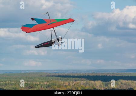 Farbenfroher roter Drachenfliegerflügel schwingt über Felder und Wälder mit blauem Himmel und Wolken im Hintergrund. Stockfoto