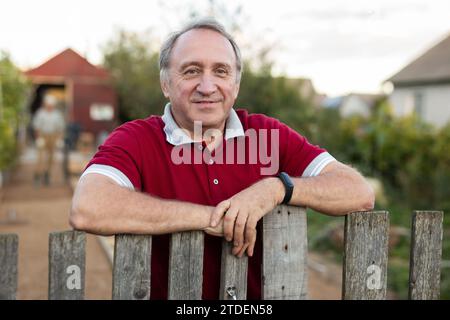Ein erwachsener Mann, der entspannt neben dem Zaun im Garten steht und lächelt Stockfoto