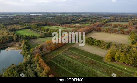 Das Bild zeigt eine weitläufige Luftaufnahme einer ländlichen Landschaft in den frühen Herbstphasen. Unten links ist ein kleiner Wasserkörper sichtbar, der den Himmel reflektiert. Die Felder zeigen verschiedene Stadien der Kultivierung und Brache, umgeben von Baumlinien, die die wechselnden Farben der Saison zeigen. In der Ferne verschmilzt die Landschaft in ein dichteres Waldgebiet, und der Horizont zeigt Hinweise auf ländliche Entwicklung. Blick aus der Vogelperspektive auf die Landschaft mit Feldern und Wasser. Hochwertige Fotos Stockfoto