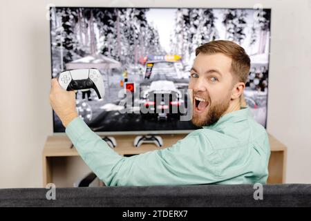 Junger Mann verbringt Zeit zu Hause, sitzt auf einer Couch im Zimmer und spielt Videospiele auf der Konsole. Erfolgreicher Mann mit Joystick zum Spielen von Actionspielen und Stockfoto