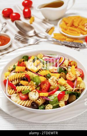Fusilli-Salat mit Mini-Mozzarella, Tomaten, Römersalat, Oliven, Zwiebeln, Paprika und Gewürze in weißer Schüssel auf weißem Holztisch mit ingre Stockfoto