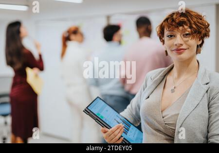 Ginger Geschäftsfrau lächelt und posiert mit dem Laptop im Büro, während ihre Kollegen hinten ein Brainstorming machen, Copy Space. Stockfoto