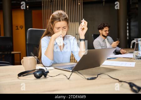 Junge kranke Geschäftsfrau, die während der Arbeit Nasentropfen in die laufende Nase aufträgt Stockfoto